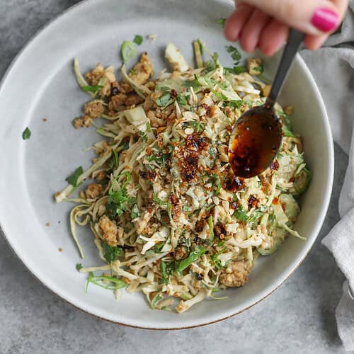 Crispy Quinoa Salad 14 A Hand With Pink Nail Polish Drizzles Sauce From A Spoon Over A Bowl Of Noodles Mixed With Ground Meat And Fresh Herbs. In The Background On A Gray Surface, A Small Bowl Of Chopped Herbs Waits Beside A Vibrant Crispy Quinoa Salad.