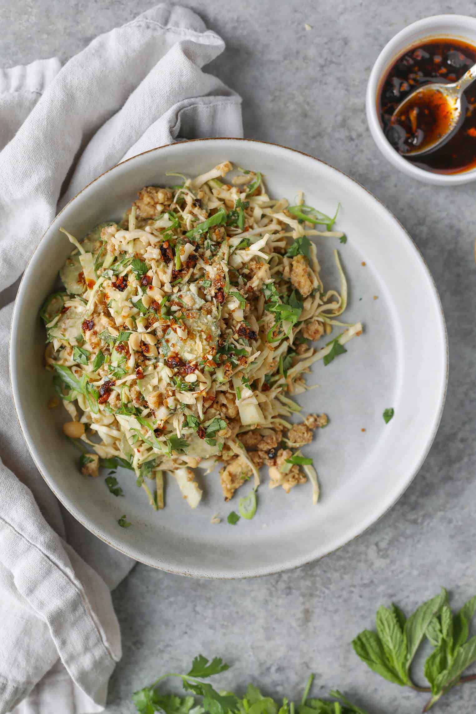 Crispy Quinoa Salad 10 A Plate Of Noodle And Crispy Quinoa Salad Topped With Herbs, Chopped Nuts, And Chili Flakes Sits On A Gray Surface Next To A Small Bowl Of Chili Sauce And A Light Gray Napkin. Fresh Cilantro And Mint Leaves Are Scattered Nearby.