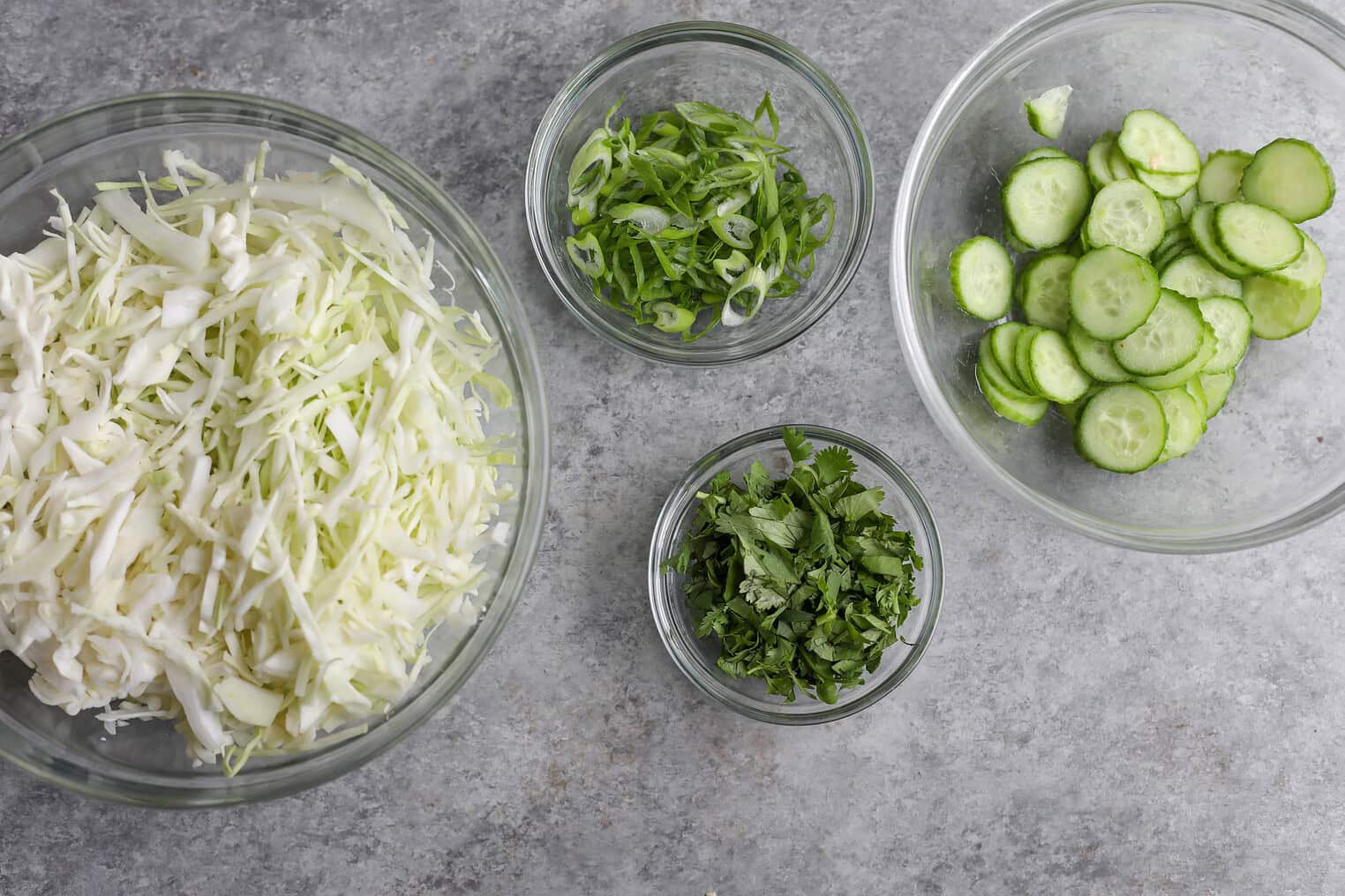 Crispy Quinoa Salad 8 Four Glass Bowls With Shredded Cabbage, Sliced Green Onions, Chopped Cilantro, And Sliced Cucumbers Are Arranged On A Gray Countertop—Perfect Toppings For A Vibrant Crispy Quinoa Salad.