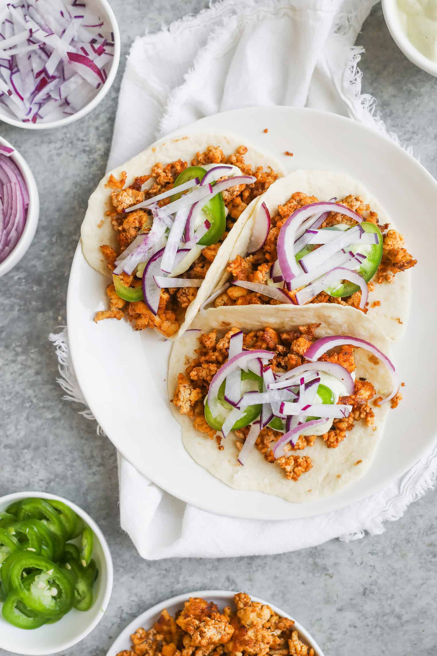 Crispy Tofu Tacos with Cashew Lime Crema 9 Three Soft Tacos Filled With Seasoned Ground Meat, Sliced JalapeƱos, And Red Onions Are Arranged On A White Plate. Bowls Of Sliced Red Onions, JalapeƱos, Extra Filling, And Crispy Tofu Tacos Surround The Plate On A Gray Surface.