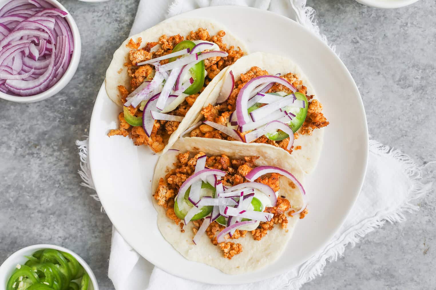 Crispy Tofu Tacos with Cashew Lime Crema 8 Three Soft Tacos Filled With Crispy Tofu, Sliced Red Onions, And JalapeƱo Peppers Are Served On A White Plate, Placed On A Light-Colored Cloth With Bowls Of Extra Toppings Nearby.