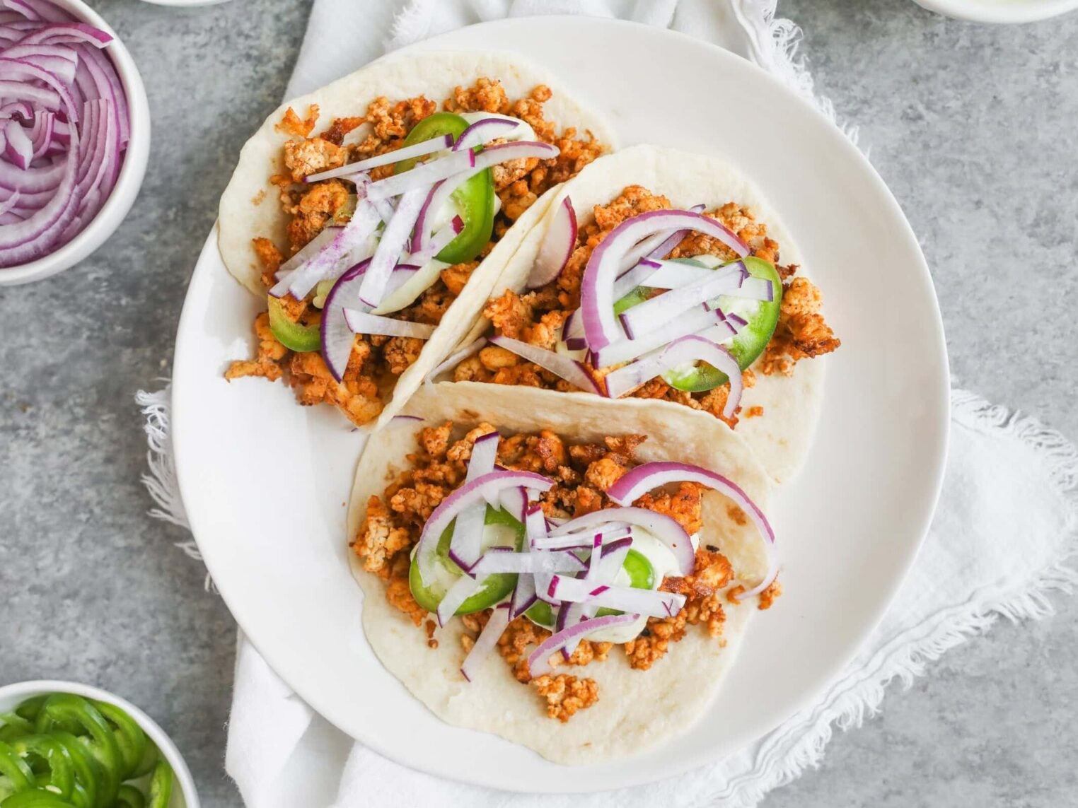 Three Soft Tacos Filled With Crispy Tofu, Sliced Red Onions, And Jalapeño Peppers Are Served On A White Plate, Placed On A Light-Colored Cloth With Bowls Of Extra Toppings Nearby.