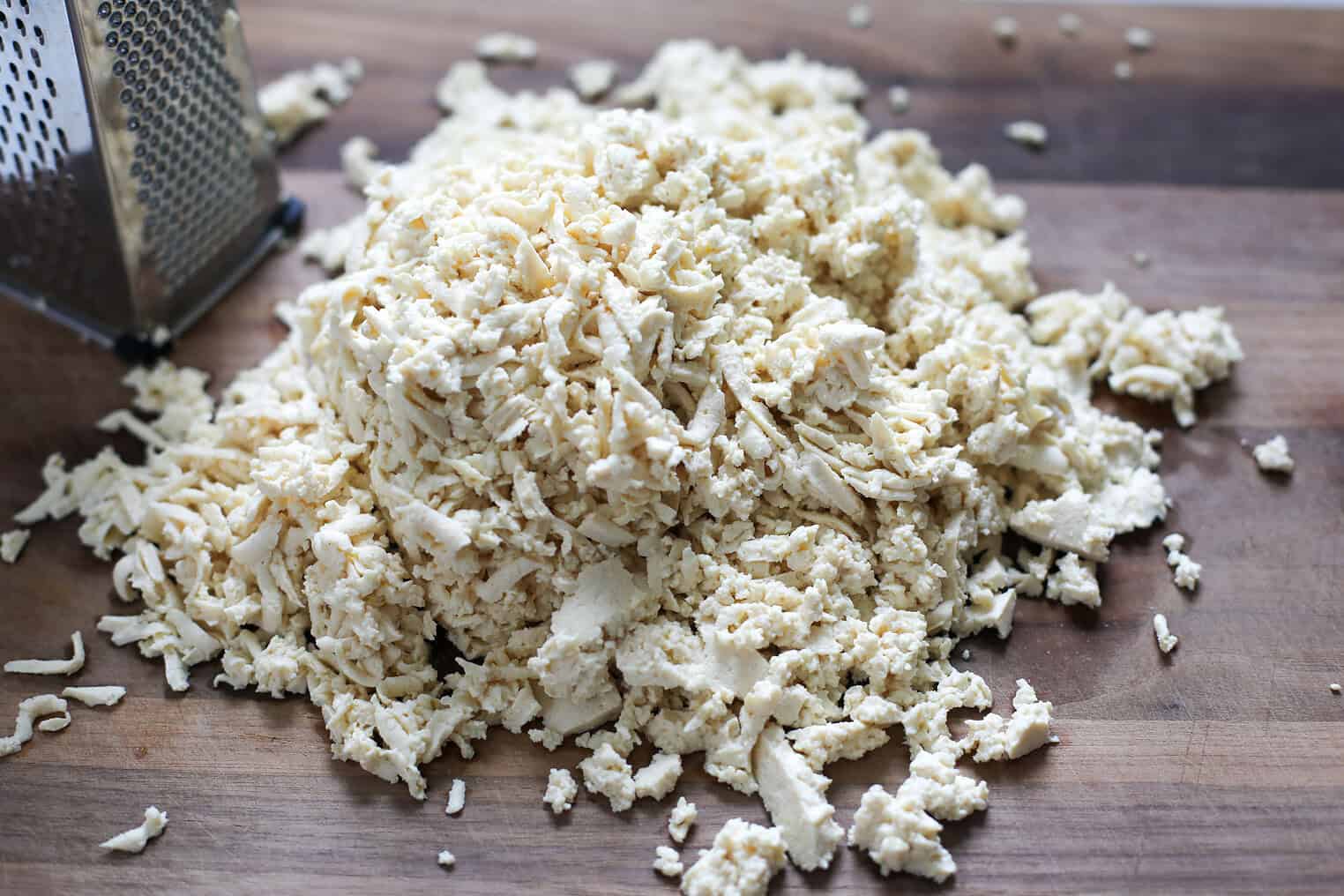 Crispy Tofu Tacos with Cashew Lime Crema 4 A Pile Of Grated Cheese Sits On A Wooden Cutting Board Next To A Metal Box Grater, Ready To Top Crispy Tofu Tacos.
