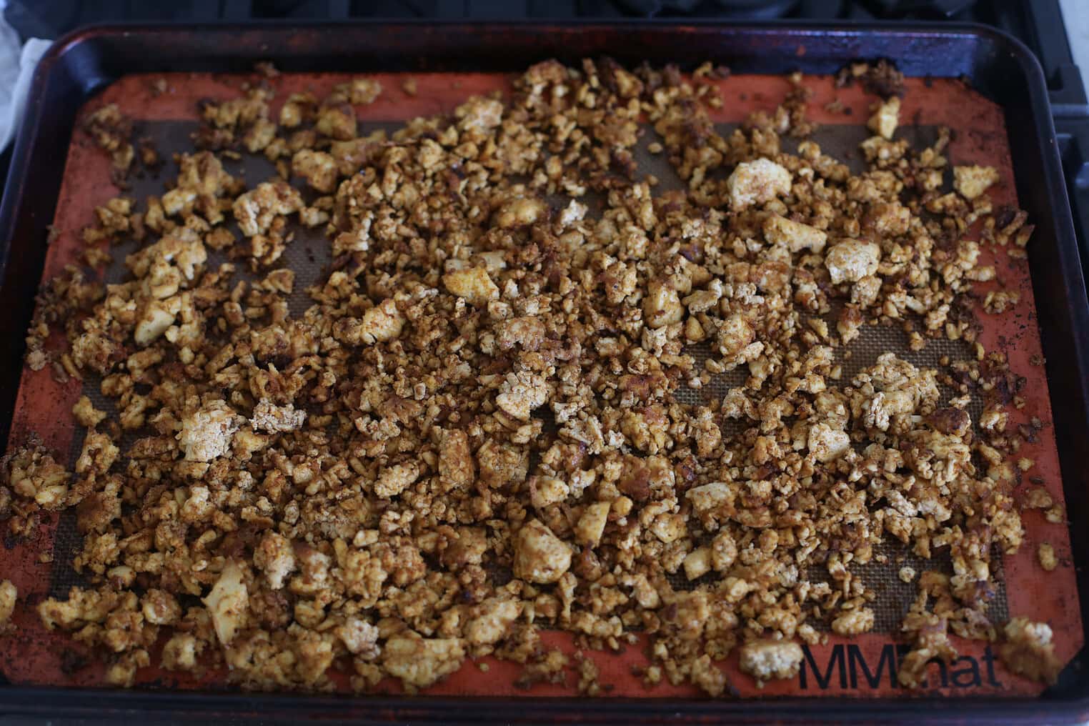 Crispy Tofu Tacos with Cashew Lime Crema 7 Crumbled, Roasted Cauliflower Spread On A Red Silicone Baking Mat Atop A Dark Baking Sheet, With Visible Seasoning And Small Browned BitsāPerfect As A Topping For Crispy Tofu Tacos. The Mat Rests On A Stove Top.