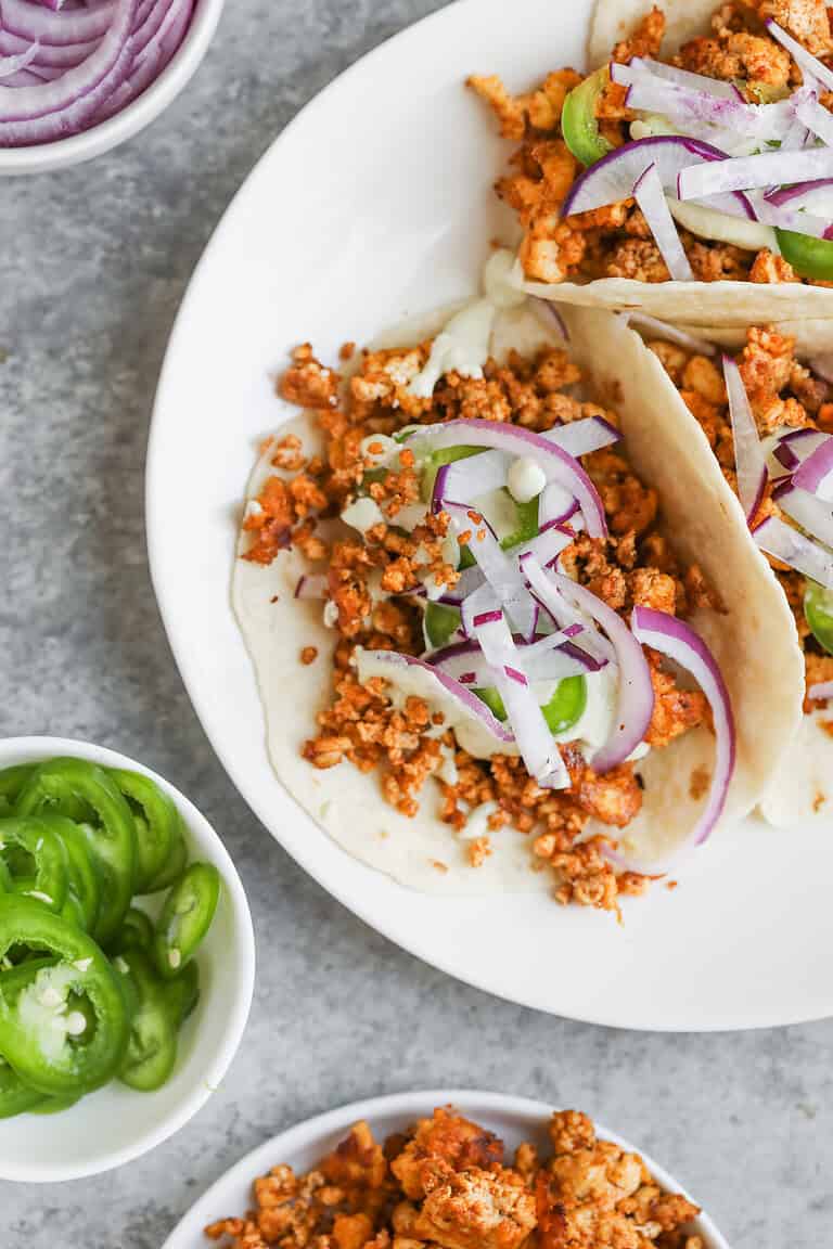 Three Crispy Tofu Tacos Filled With Seasoned Crumbled Tofu, Sliced Red Onions, And Green Bell Peppers On A White Plate. Nearby Are Small Bowls With Sliced Jalapeños, Extra Tofu, And More Red Onions For Topping.