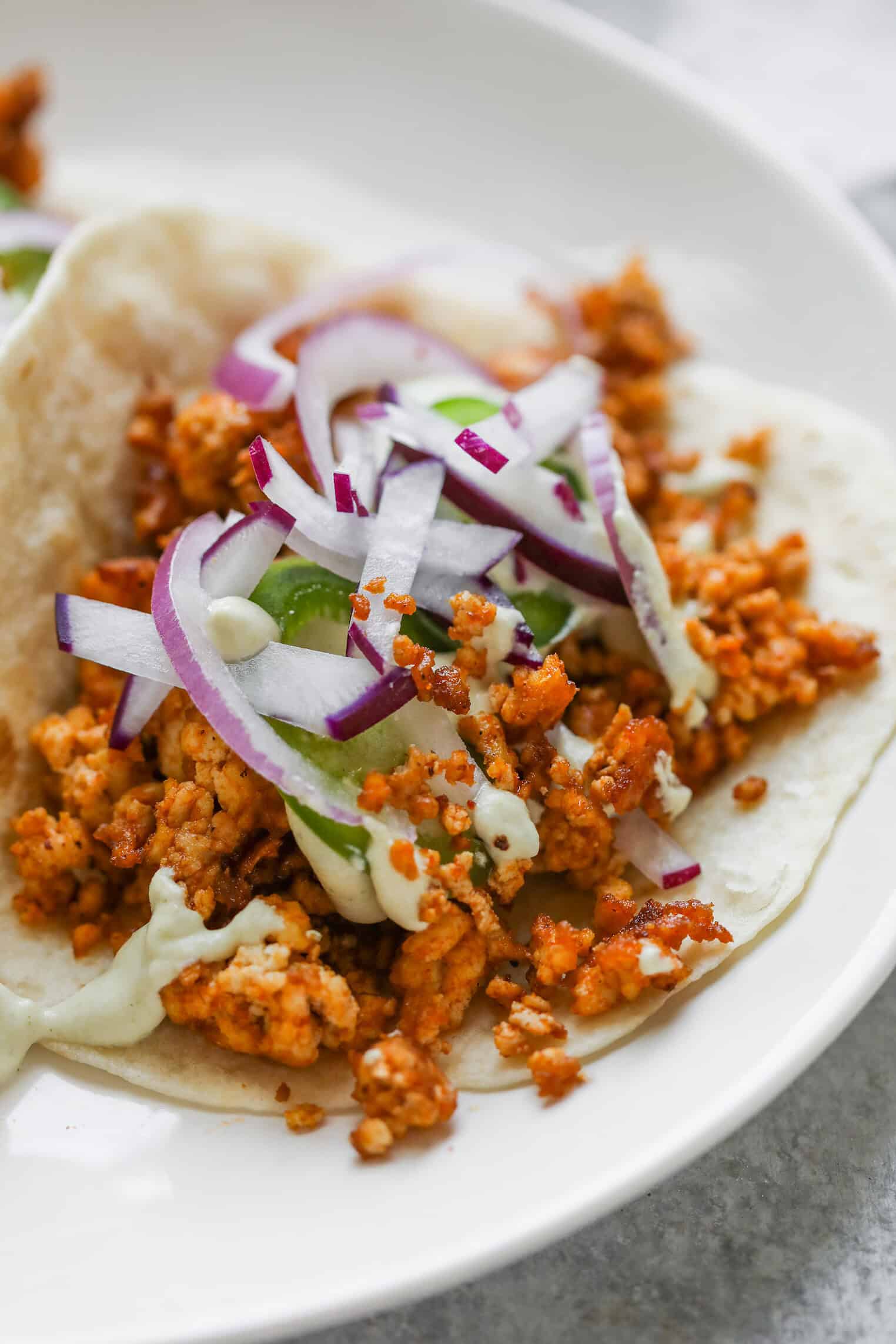 Crispy Tofu Tacos with Cashew Lime Crema 2 A Close-Up Of Crispy Tofu Tacos Filled With Crumbled Spiced Tofu, Sliced Red Onions, JalapeƱo Slices, Radish Strips, And A Drizzle Of Creamy Sauce On A White Plate.