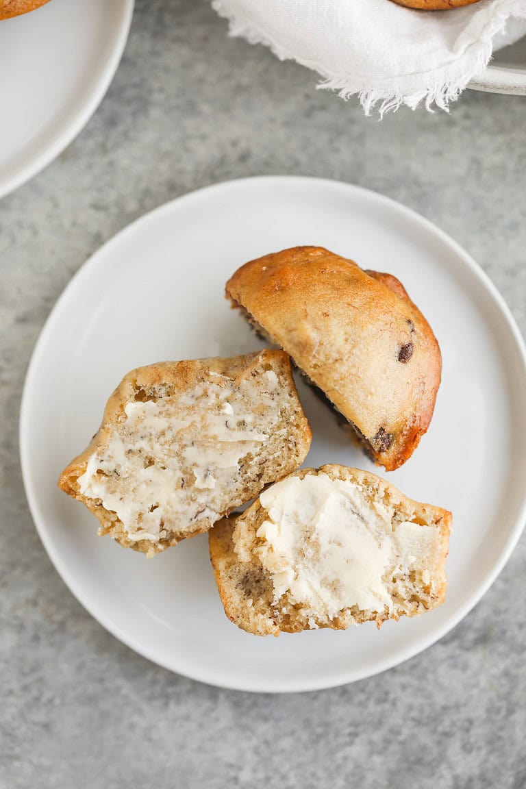 A Split Vegan Banana Muffin On A White Plate. One Half Is Spread With Butter, And The Other Is Positioned Top Down, Showcasing Its Golden-Brown, Slightly Speckled Crust. The Background Features A Gray, Textured Surface.