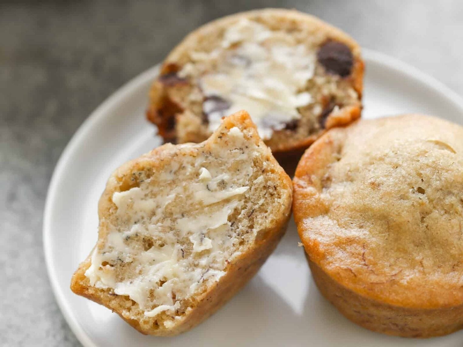 Vegan Banana Recipes 2 Three Vegan Banana Muffins Rest On A White Plate. One Muffin Is Whole, While The Other Two Are Halved, Revealing Their Moist Texture. One Of The Halves Is Smeared With Butter. A Bowl With More Delectable Muffins Sits In The Background, Inviting You To Indulge In A Plant-Based Treat.