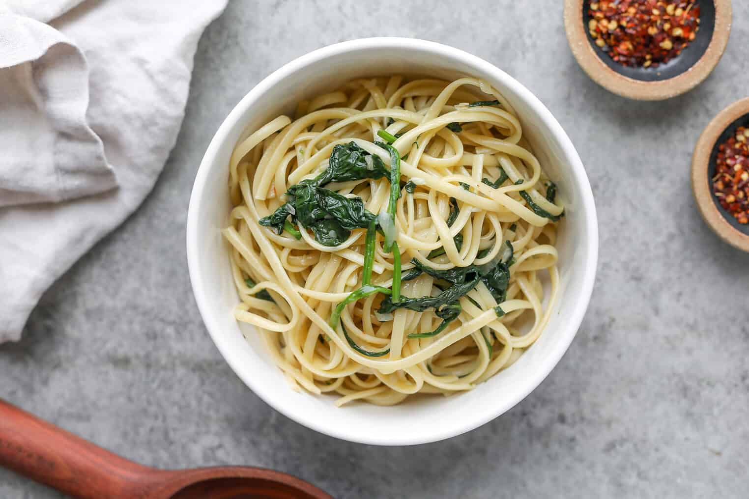 Easy Vegan Pasta 7 A White Bowl Filled With Vegan Pasta Linguine And Wilted Spinach Sits On A Gray Surface, With Small Bowls Of Red Pepper Flakes And A White Napkin Nearby.