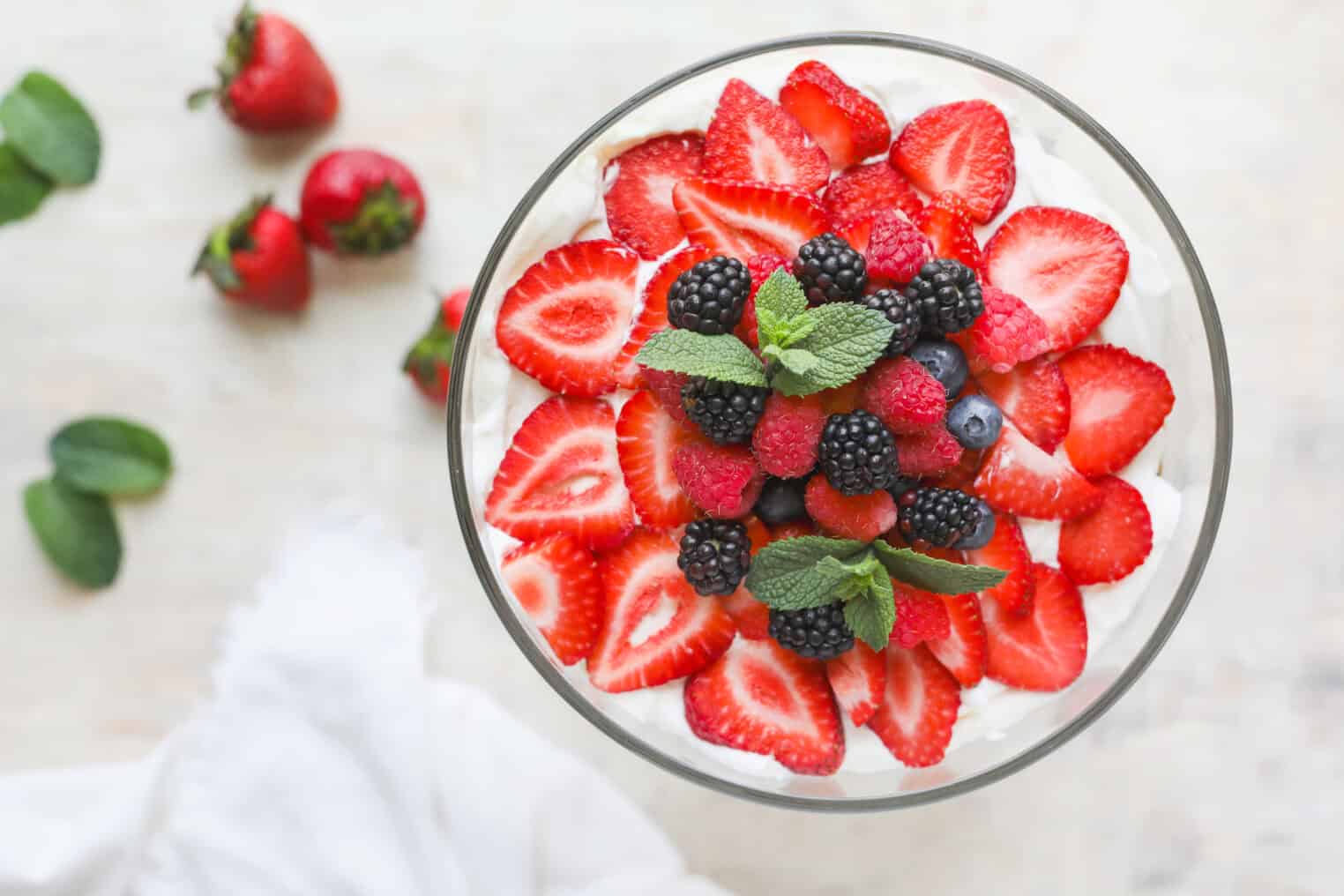 Fresh Berry Trifle 4 A Glass Bowl Filled With Whipped Cream And Topped With Sliced Strawberries, Blackberries, Raspberries, Blueberries, And Fresh Mint Evokes A Classic Berry Trifle. Whole Strawberries And Mint Are Scattered Nearby On A Light Surface.