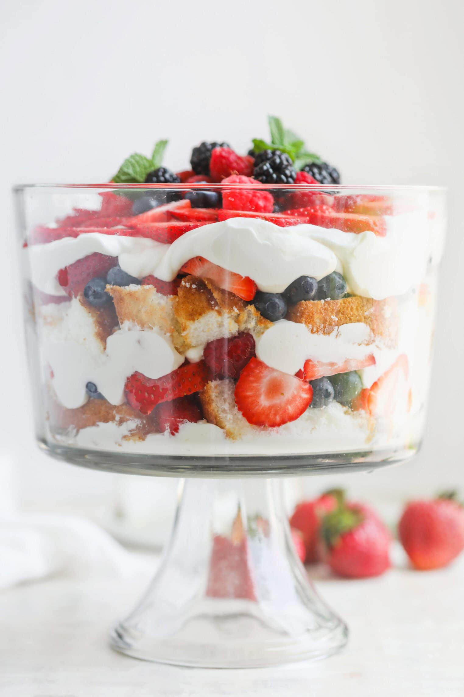 Fresh Berry Trifle 1 A Glass Trifle Bowl Filled With Layers Of Whipped Cream, Sponge Cake, Strawberries, Blueberries, And Blackberries Creates A Stunning Berry Trifle, Garnished With Fresh Berries And Mint. Two Strawberries Sit Beside The Bowl.