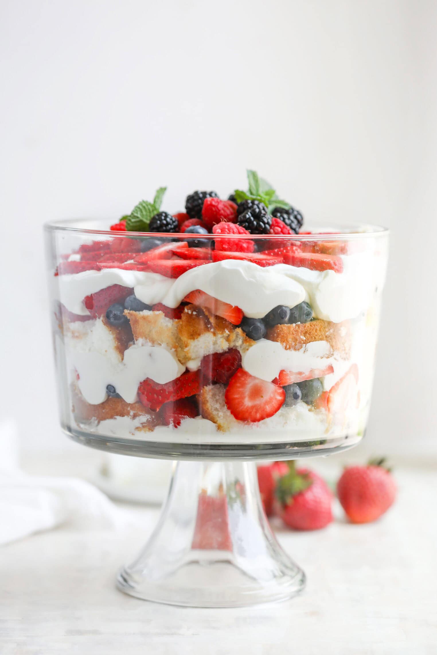 Fresh Berry Trifle 11 A Glass Trifle Dish Filled With Layers Of Cake, Whipped Cream, And Fresh Berries Creates A Stunning Berry Trifle, Garnished With Mint Leaves On Top. Two Strawberries Rest On The Surface Beside The Dish.