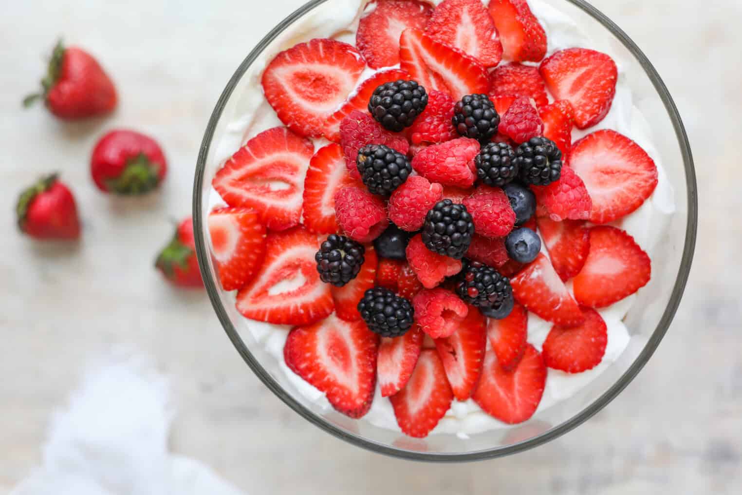 Fresh Berry Trifle 10 A Glass Bowl Filled With Whipped Cream, Topped With Sliced Strawberries, Blackberries, Raspberries, And Blueberries Resembles A Delightful Berry Trifle; Whole Strawberries Are Scattered On The Light-Colored Surface Beside The Bowl.