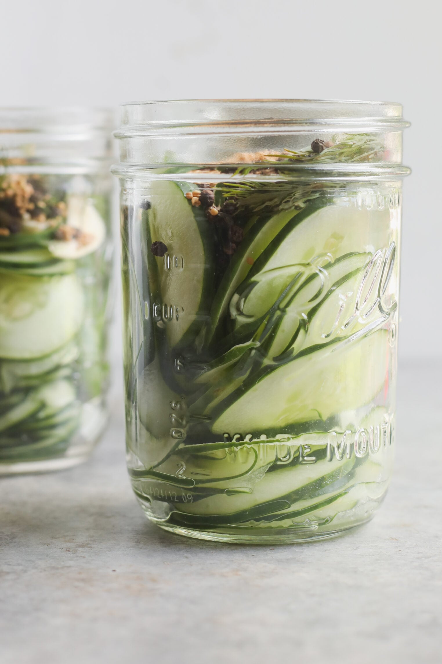 A Clear Glass Mason Jar Filled With Sliced Cucumbers And Herbs For Refrigerator Pickles Sits On A Light-Colored Surface, With Another Similar Jar Partially Visible In The Background.