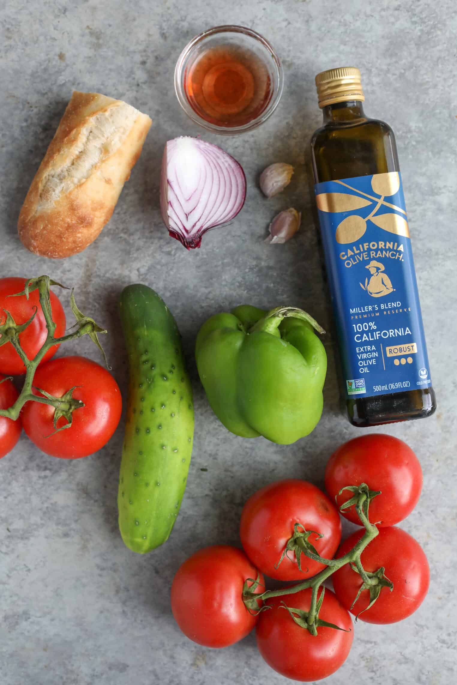 Gazpacho 3 A Bottle Of California Olive Ranch Olive Oil, A Small Baguette, Half A Red Onion, Two Garlic Cloves, A Small Glass Of Vinegar, A Green Bell Pepper, A Cucumber, And Fresh Tomatoes On A Gray Surface.