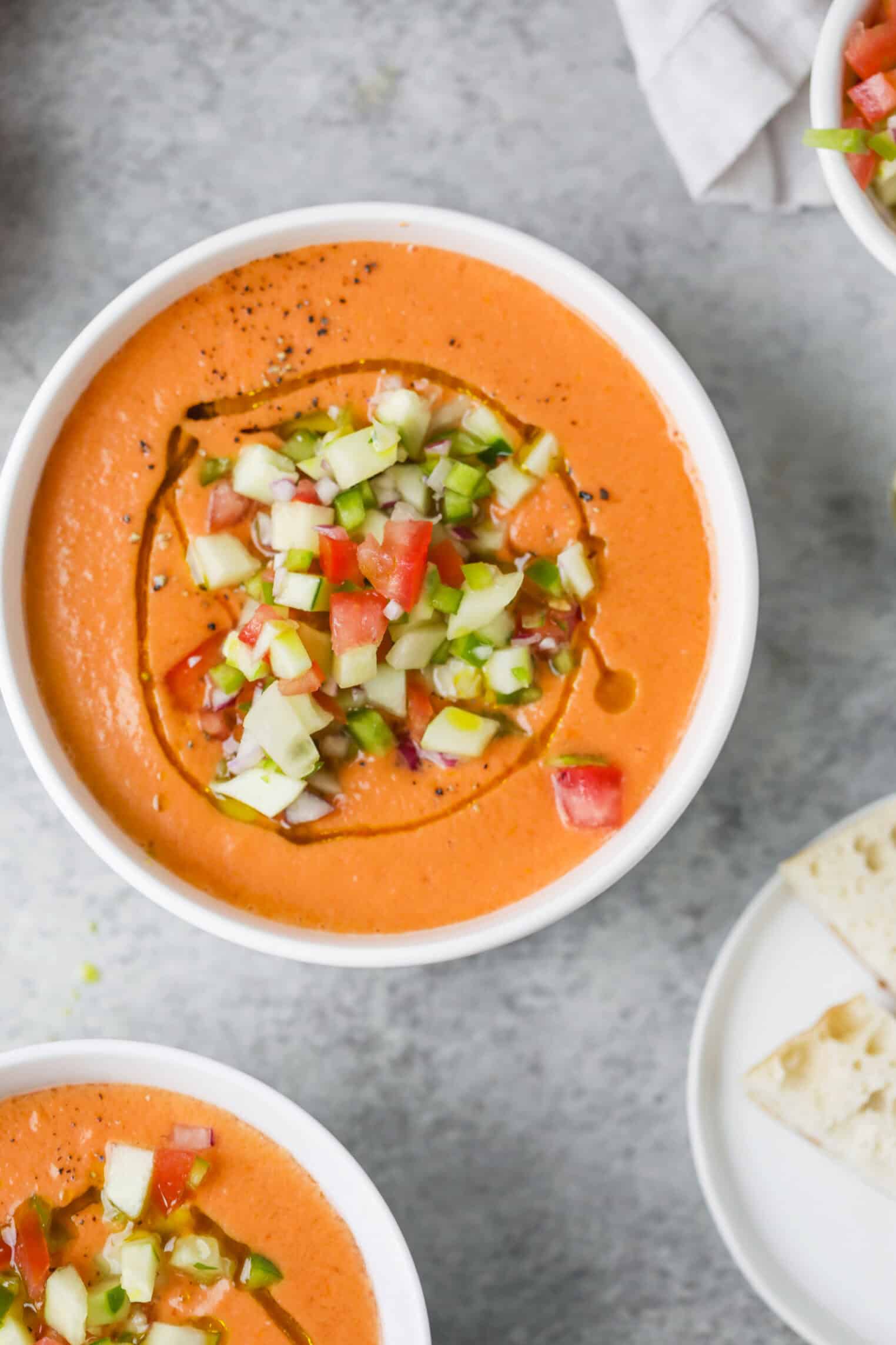 Gazpacho 1 A Bowl Of Creamy Orange Soup Topped With Diced Cucumber, Tomato, And Onion, Drizzled With Olive Oil. The Bowl Sits On A Light Surface Next To A Plate With Pieces Of Bread.
