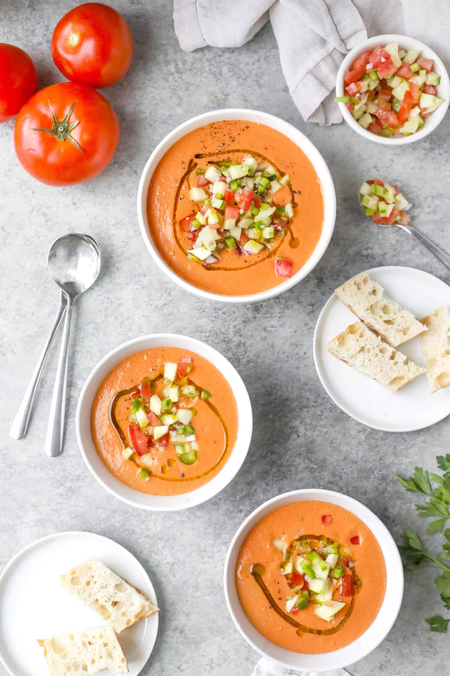 Gazpacho 4 Four Bowls Of Creamy Gazpacho Garnished With Diced Cucumber And Tomato, Drizzled With Olive Oil. Slices Of Bread, Fresh Tomatoes, Spoons, And A Napkin Are Arranged On A Light Gray Surface.