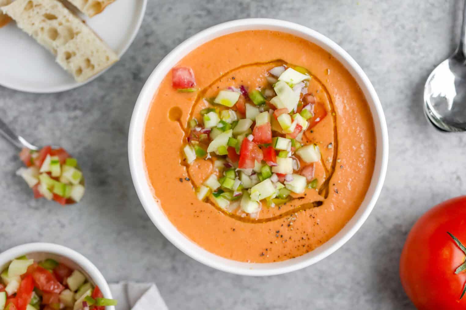 Gazpacho 7 A Bowl Of Creamy Gazpacho Soup Garnished With Diced Cucumber And Tomato, Drizzled With Olive Oil, Sits On A Grey Surface. Nearby Are A Spoon, A Plate With Bread, And A Fresh Tomato Ready To Complement The Gazpacho.