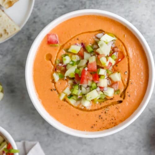 Gazpacho 10 A Bowl Of Creamy Gazpacho Soup Garnished With Diced Cucumber And Tomato, Drizzled With Olive Oil, Sits On A Grey Surface. Nearby Are A Spoon, A Plate With Bread, And A Fresh Tomato Ready To Complement The Gazpacho.