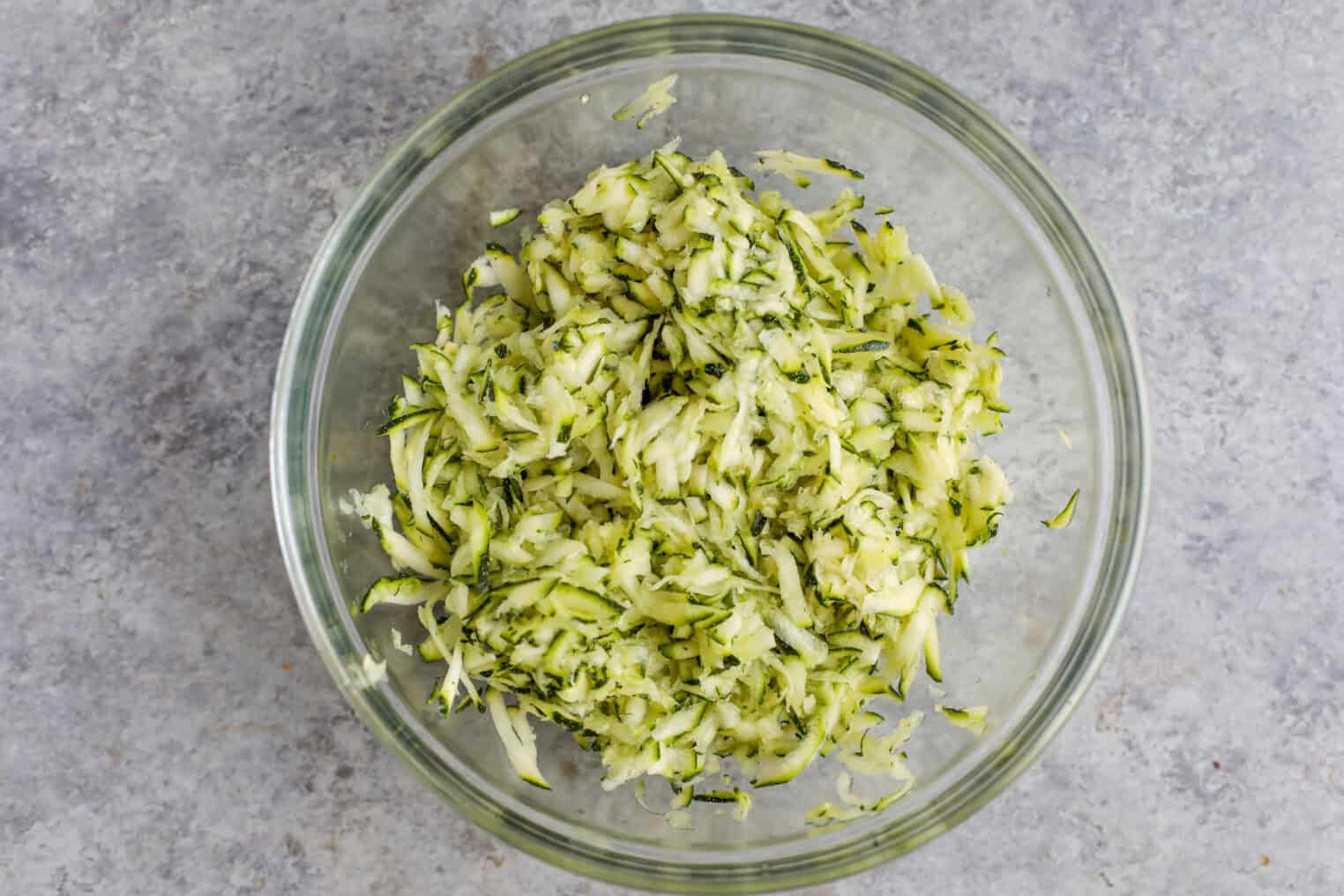 Greek Quinoa Zucchini Fritters 5 A Clear Glass Bowl Filled With Freshly Grated Zucchini Sits On A Gray, Textured Surface—Perfect For Making Quinoa Zucchini Fritters. The Finely Shredded Mix Shows Light Green And White Pieces With Flecks Of Darker Green Skin.
