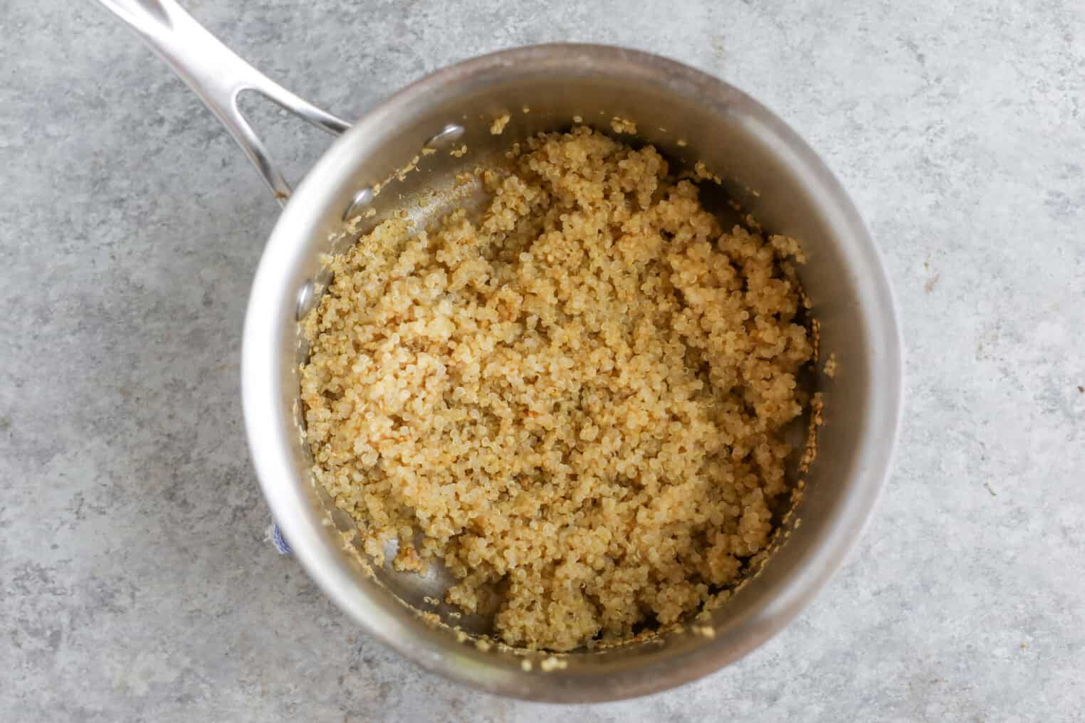 Greek Quinoa Zucchini Fritters 4 A Stainless Steel Pot Filled With Cooked Quinoa Sits On A Gray Textured Surface, Perfect As A Base For Making Quinoa Zucchini Fritters. The Quinoa Looks Fluffy And Slightly Golden In Color.