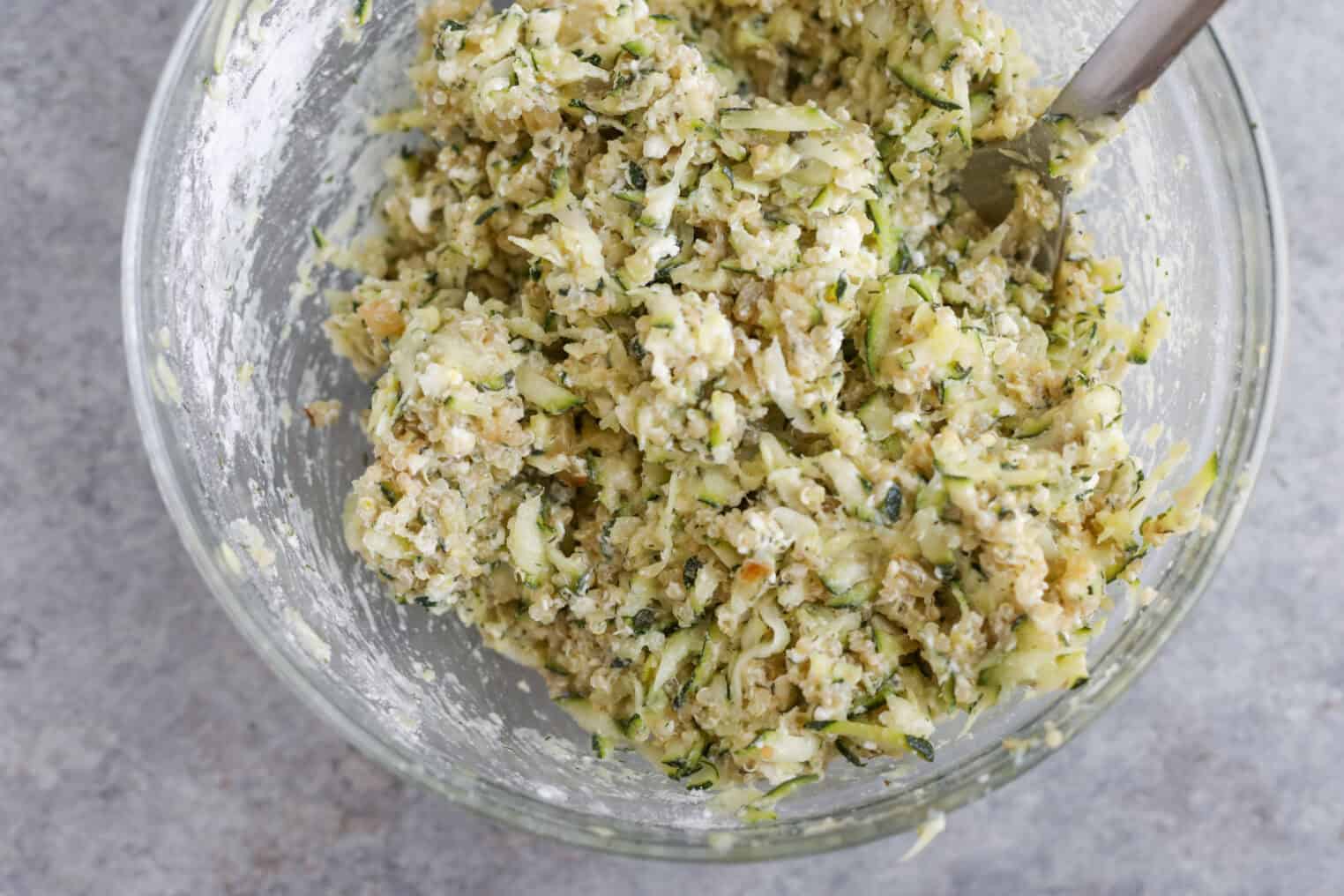 Greek Quinoa Zucchini Fritters 7 A Clear Glass Bowl Filled With A Mixture For Quinoa Zucchini Fritters—Shredded Zucchini, Flour, And Seasonings—Being Stirred With A Metal Spoon On A Gray Countertop.