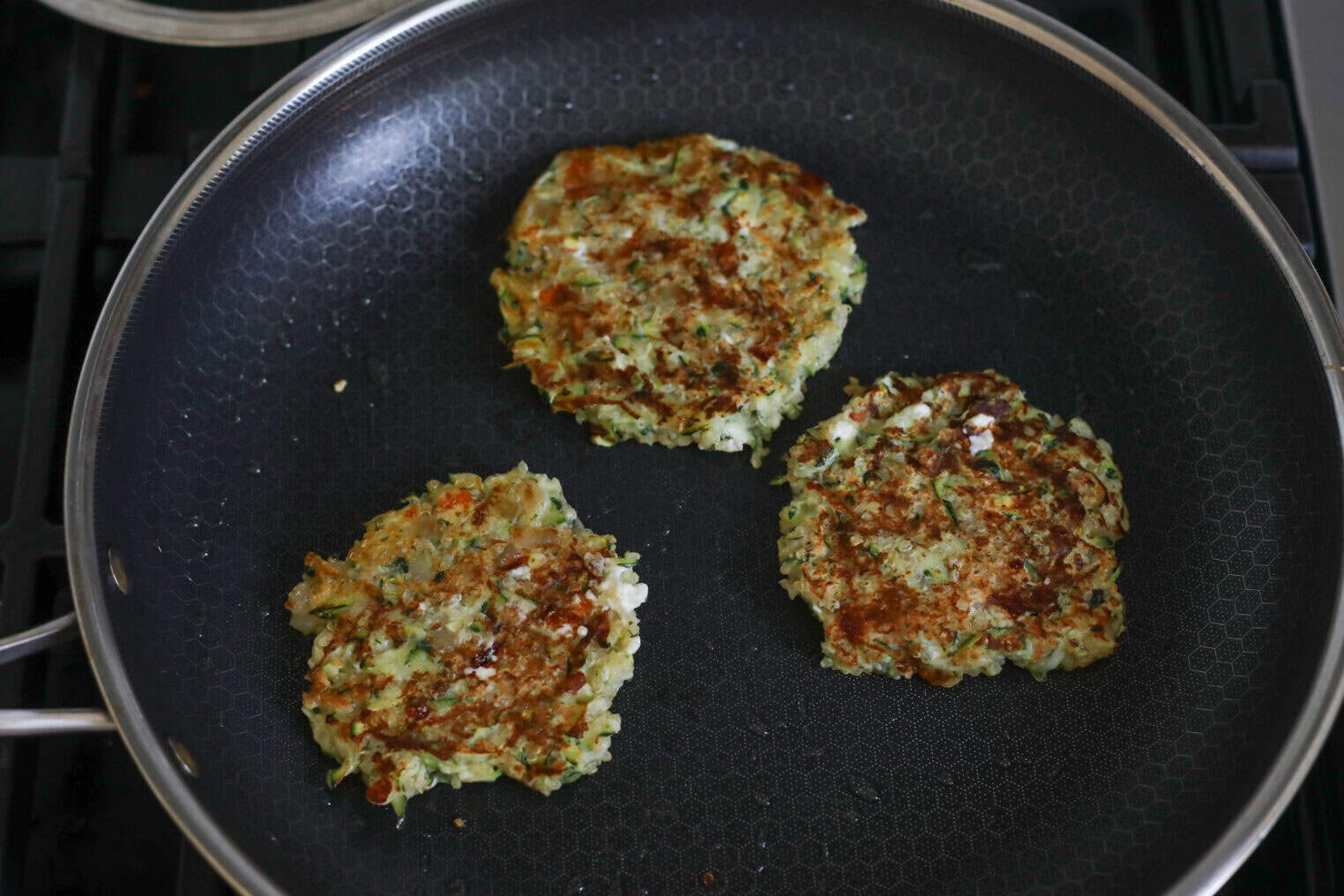 Greek Quinoa Zucchini Fritters 8 Three Golden-Brown Quinoa Zucchini Fritters Are Cooking In A Non-Stick Frying Pan On The Stove. The Patties Have A Slightly Rough Texture And Are Speckled With Green And White Ingredients.