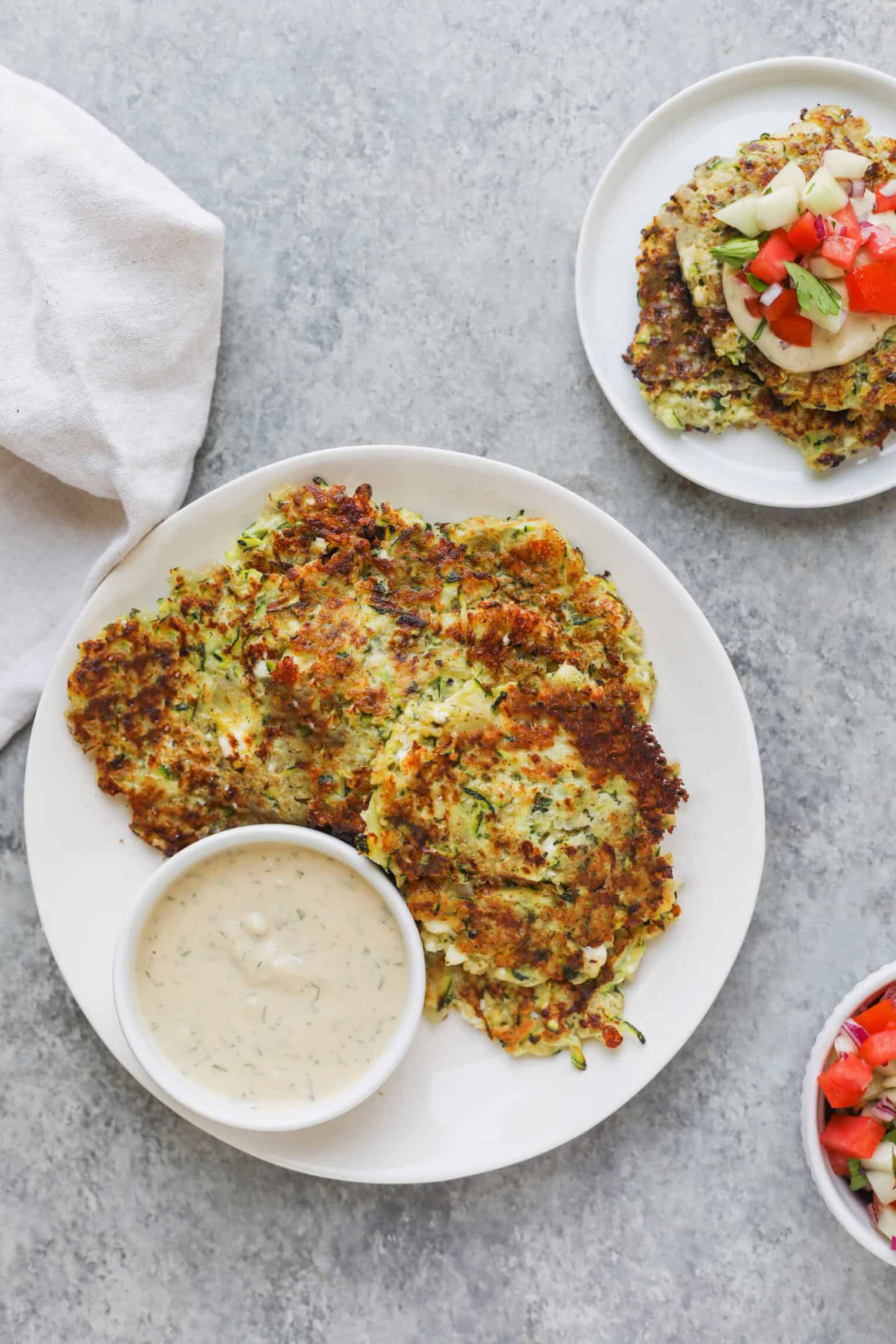 Greek Quinoa Zucchini Fritters 3 Three Golden-Brown Quinoa Zucchini Fritters On A White Plate With A Bowl Of Creamy Dipping Sauce, Set On A Gray Surface. A Small Plate With More Fritters And A Bowl Of Diced Vegetables Are Nearby.