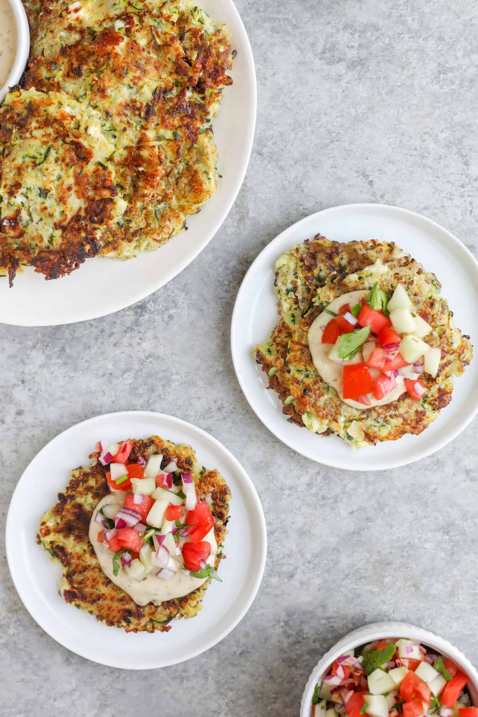 Greek Quinoa Zucchini Fritters 9 Two Small White Plates And One Large Plate Display Golden-Brown Quinoa Zucchini Fritters. Some Fritters Are Topped With Diced Tomato, Cucumber, Red Onion, And Herbs. A Small Bowl Of Similar Chopped Vegetable Topping Sits Nearby.