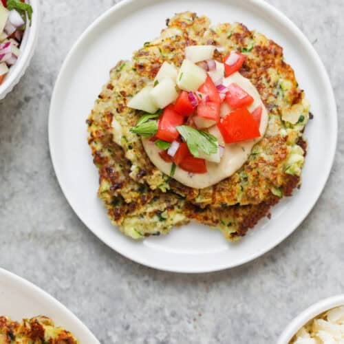 Greek Quinoa Zucchini Fritters 12 A Plate With Two Golden-Brown Quinoa Zucchini Fritters Topped With Diced Tomato, Cucumber, And Fresh Herbs. Surrounding Bowls Contain More Fritters, Chopped Salad, And Crumbled Cheese On A Light Gray Surface.