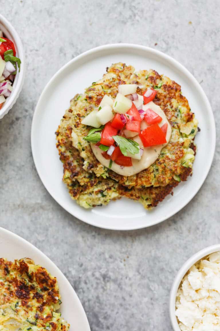 A Plate With Two Golden-Brown Quinoa Zucchini Fritters Topped With Diced Tomato, Cucumber, And Fresh Herbs. Surrounding Bowls Contain More Fritters, Chopped Salad, And Crumbled Cheese On A Light Gray Surface.