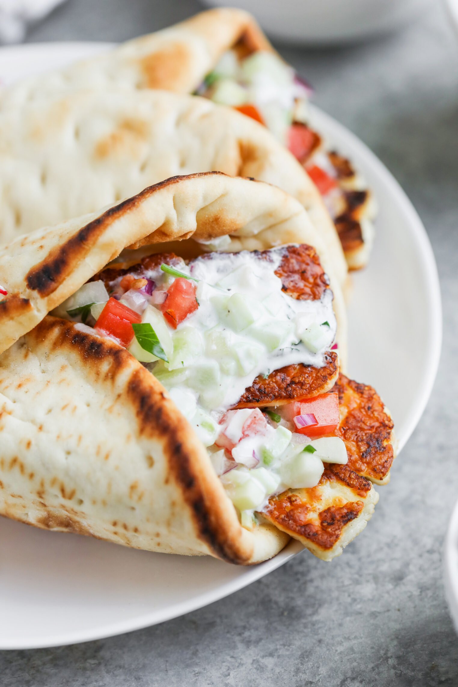 Halloumi Gyros 1 Two Halloumi Gyros Pita Wraps Filled With Grilled Halloumi Cheese, Chopped Tomatoes, Cucumbers, Red Onions, And Creamy Tzatziki Sauce Are Served On A White Plate.