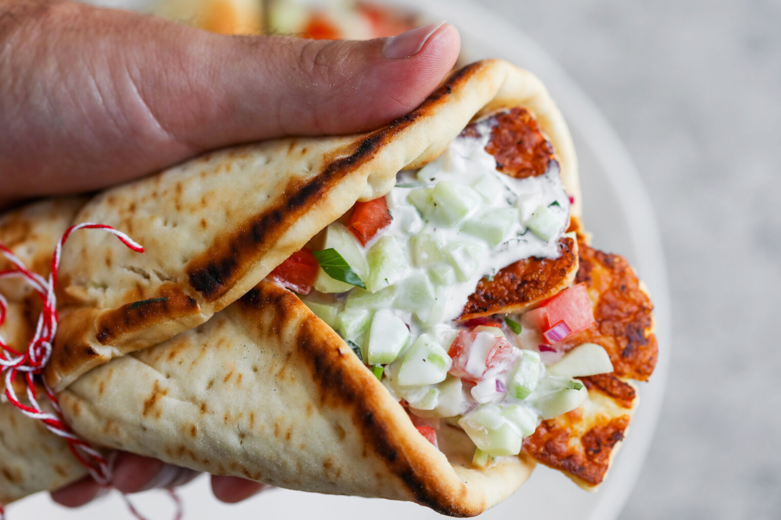 Halloumi Gyros 6 A Hand Holding A Grilled Pita Wrap Filled With Sliced Halloumi Cheese, Diced Tomatoes, Cucumbers, Onions, And Creamy Tzatziki Sauce, Tied With A Red And White String—An Irresistible Halloumi Gyros Delight.