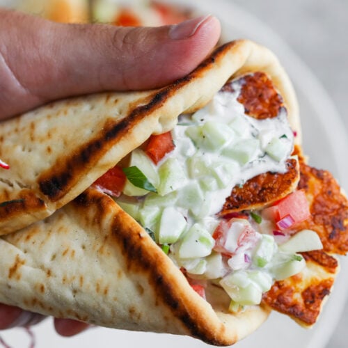 Halloumi Gyros 9 A Hand Holding A Grilled Pita Wrap Filled With Sliced Halloumi Cheese, Diced Tomatoes, Cucumbers, Onions, And Creamy Tzatziki Sauce, Tied With A Red And White String—An Irresistible Halloumi Gyros Delight.