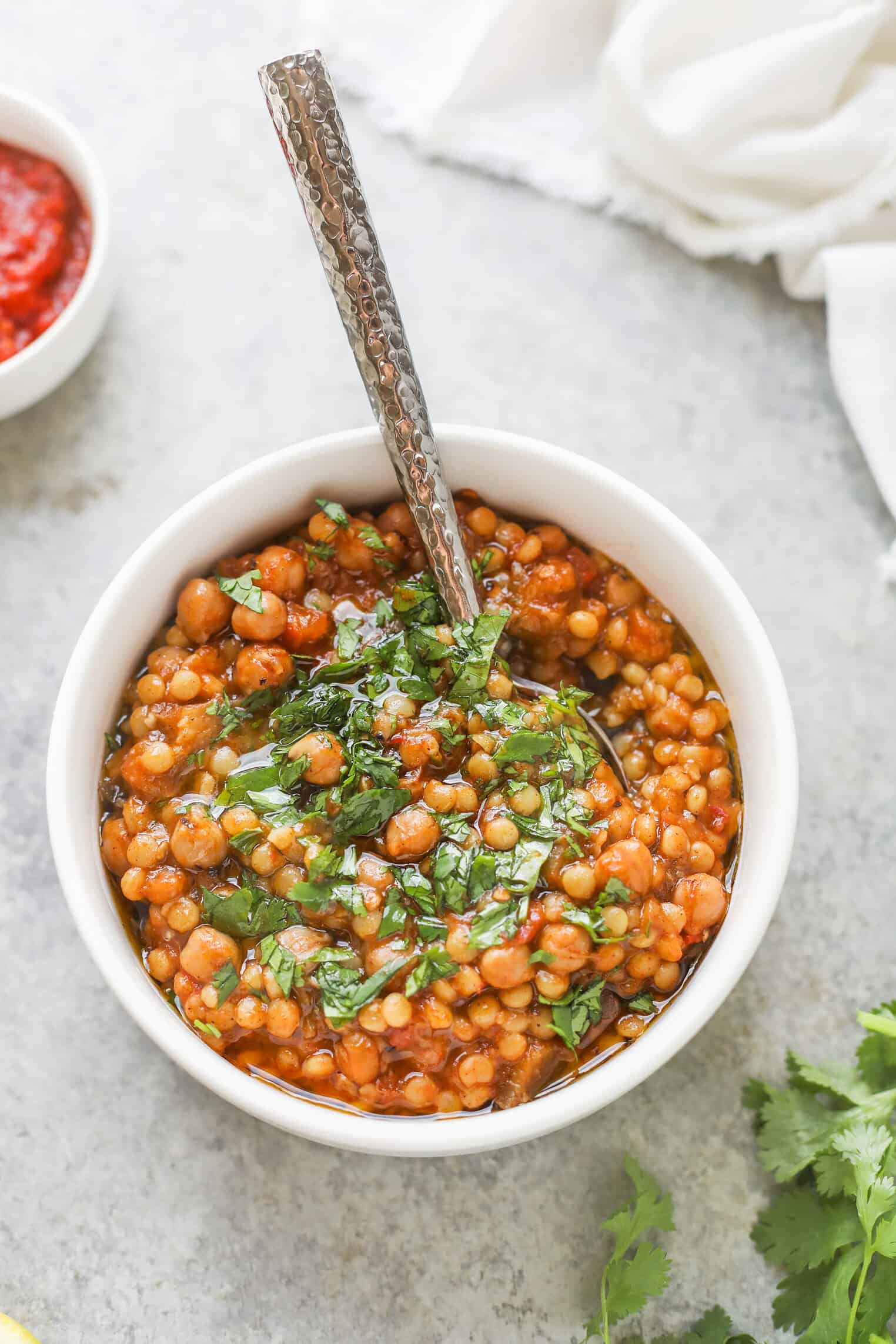 A White Bowl Filled With Red Lentil Stew And Garnished With Chopped Fresh Cilantro Sits On A Light Gray Surface, Accompanied By A Spoon, A Small Dish Of Red Sauce, Harissa Couscous, And Sprigs Of Cilantro Nearby.