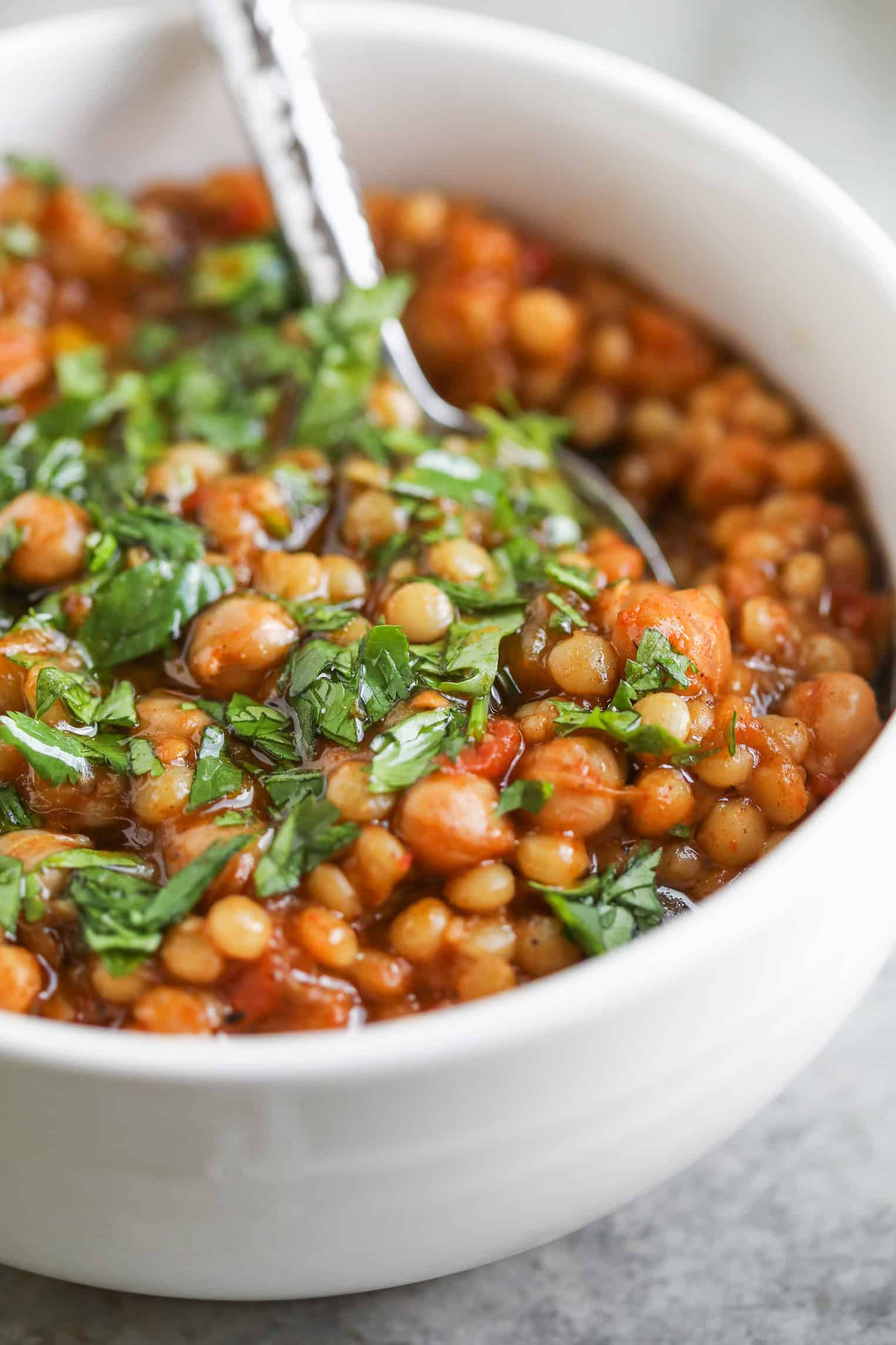 A Close-Up Of A White Bowl Filled With Lentil Stew And Harissa Couscous, Topped With Chopped Fresh Herbs. A Spoon Is Resting Inside The Bowl. The Stew Appears Thick And Hearty, With Visible Tomatoes And Herbs.