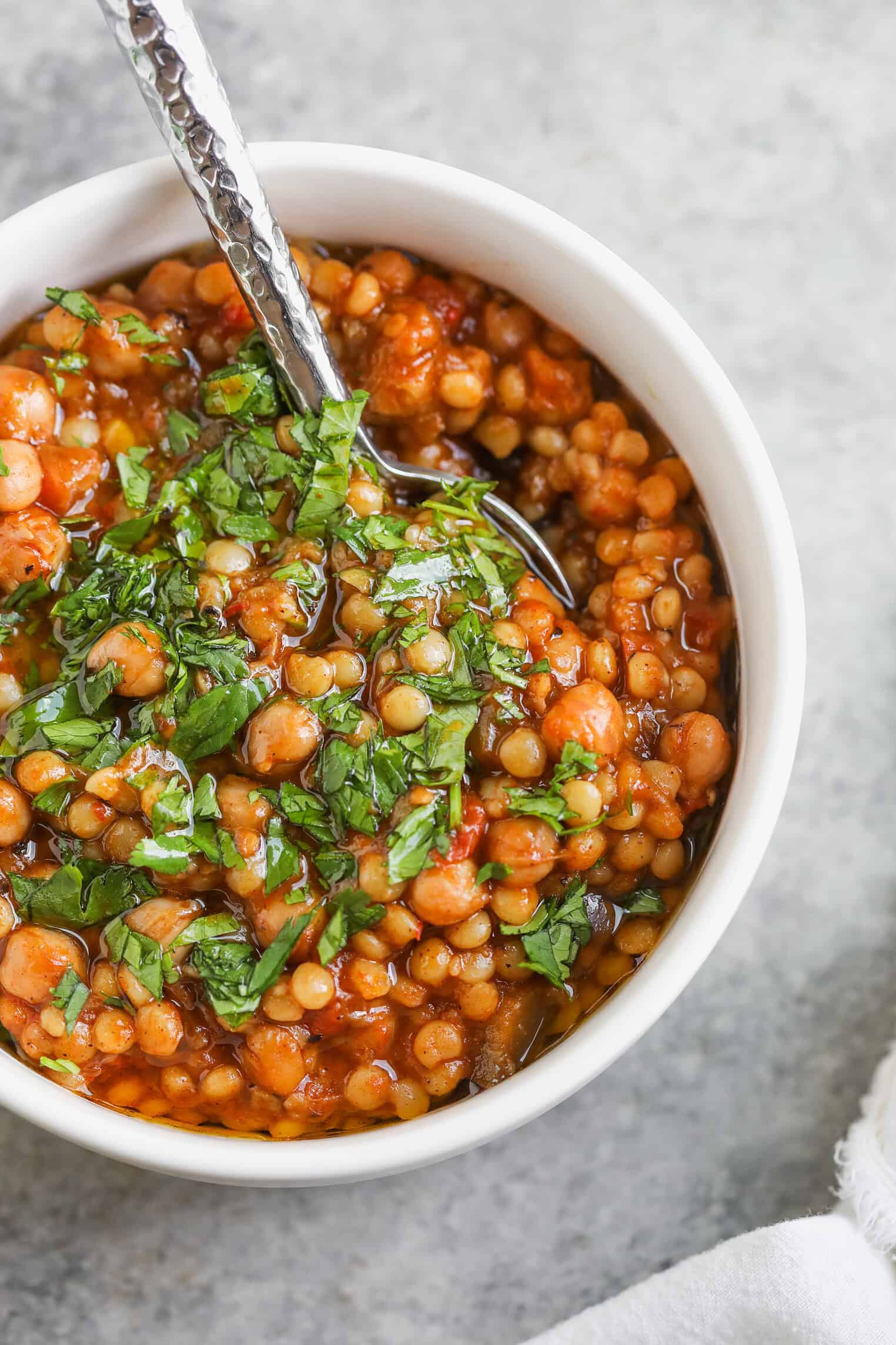 A White Bowl Filled With Tomato-Based Harissa Couscous Stew, Garnished With Chopped Fresh Herbs, With A Spoon Resting Inside. The Bowl Sits On A Light Gray Surface.