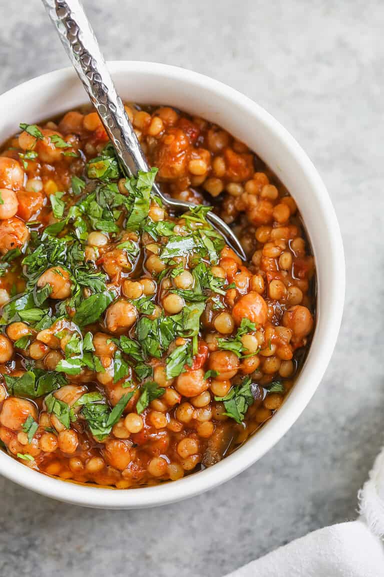 A White Bowl Filled With Tomato-Based Harissa Couscous Stew, Garnished With Chopped Fresh Herbs, With A Spoon Resting Inside. The Bowl Sits On A Light Gray Surface.