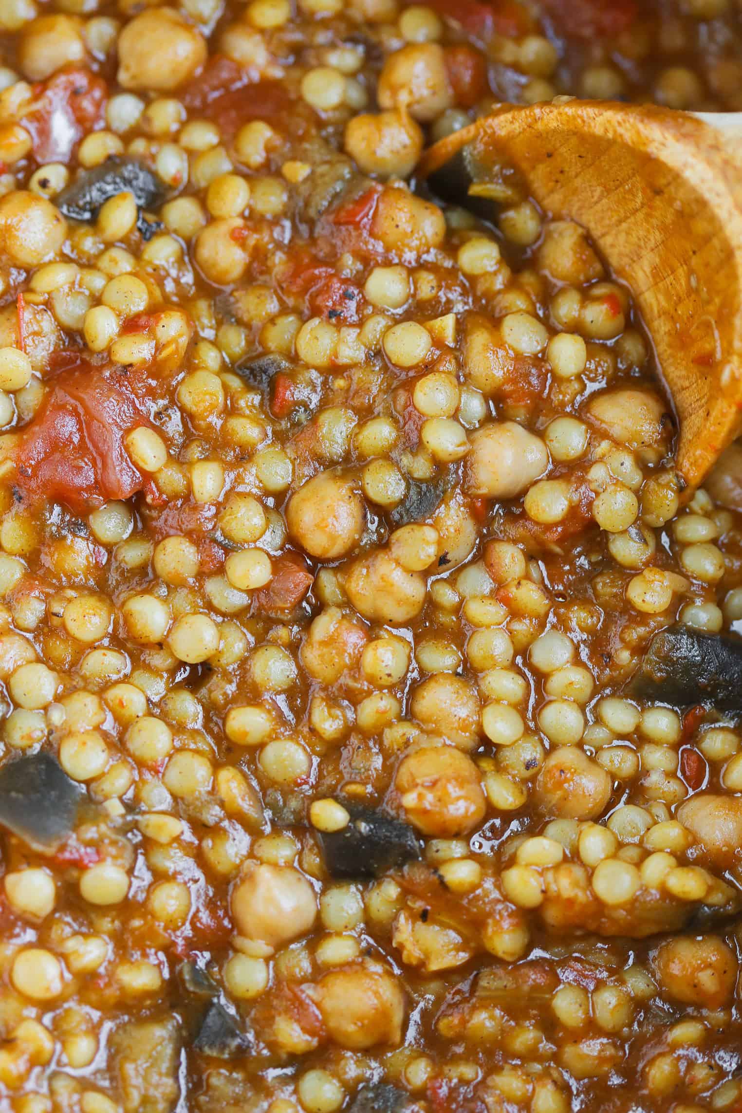 Close-Up Of A Thick Stew With Chickpeas, Lentils, Tomato Chunks, And Eggplant, Being Stirred With A Wooden Spoon. Rich And Hearty, This Reddish-Brown Mixture Pairs Perfectly With Harissa Couscous For Extra Flavor And Warmth.