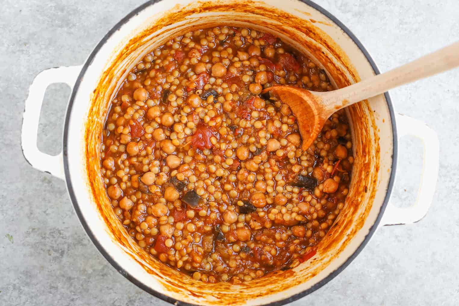 A White Pot Filled With A Thick, Hearty Stew Or Soup Containing Chickpeas, Lentils, Tomatoes, Spices, And A Hint Of Harissa Couscous. A Wooden Spoon Rests Inside The Pot, Mixing The Flavorful Ingredients.