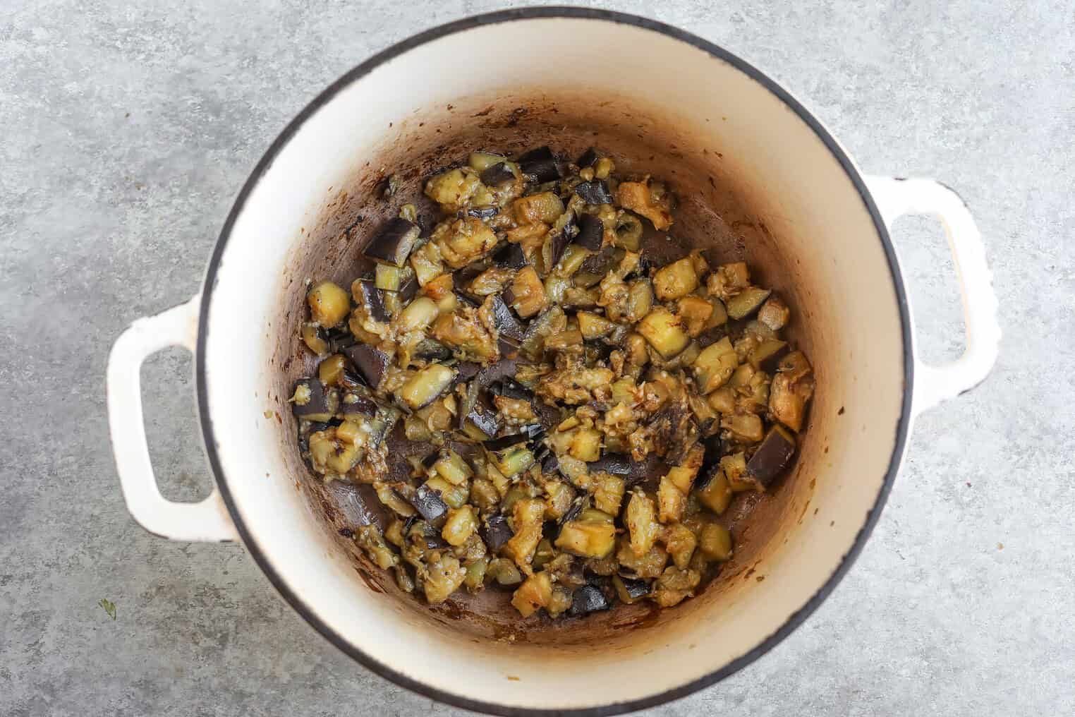 A White Pot With Cooked, Diced Eggplant Pieces, Slightly Browned And Mixed With Seasonings, Sits On A Light Gray Countertop—Perfect For Serving Over Harissa Couscous.