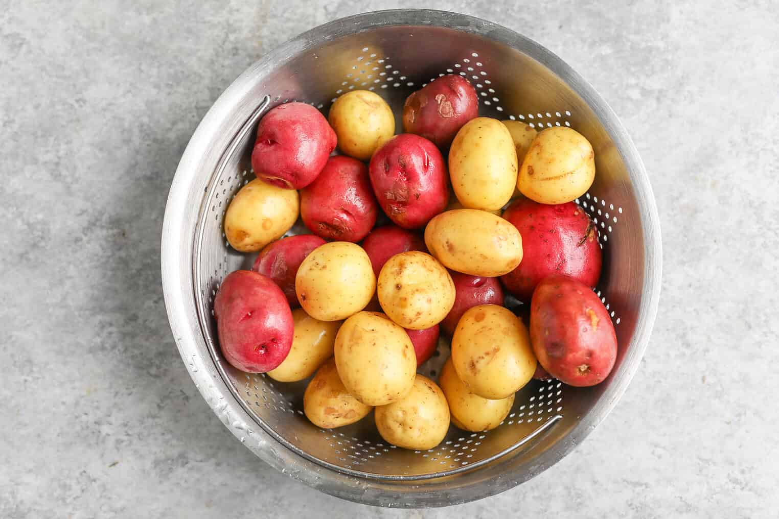 Creamy Herbed Potato Salad 4 A Metal Colander Filled With Small Red And Yellow Potatoes, Perfect For Making Herbed Potato Salad, Sits On A Light Gray Textured Surface.