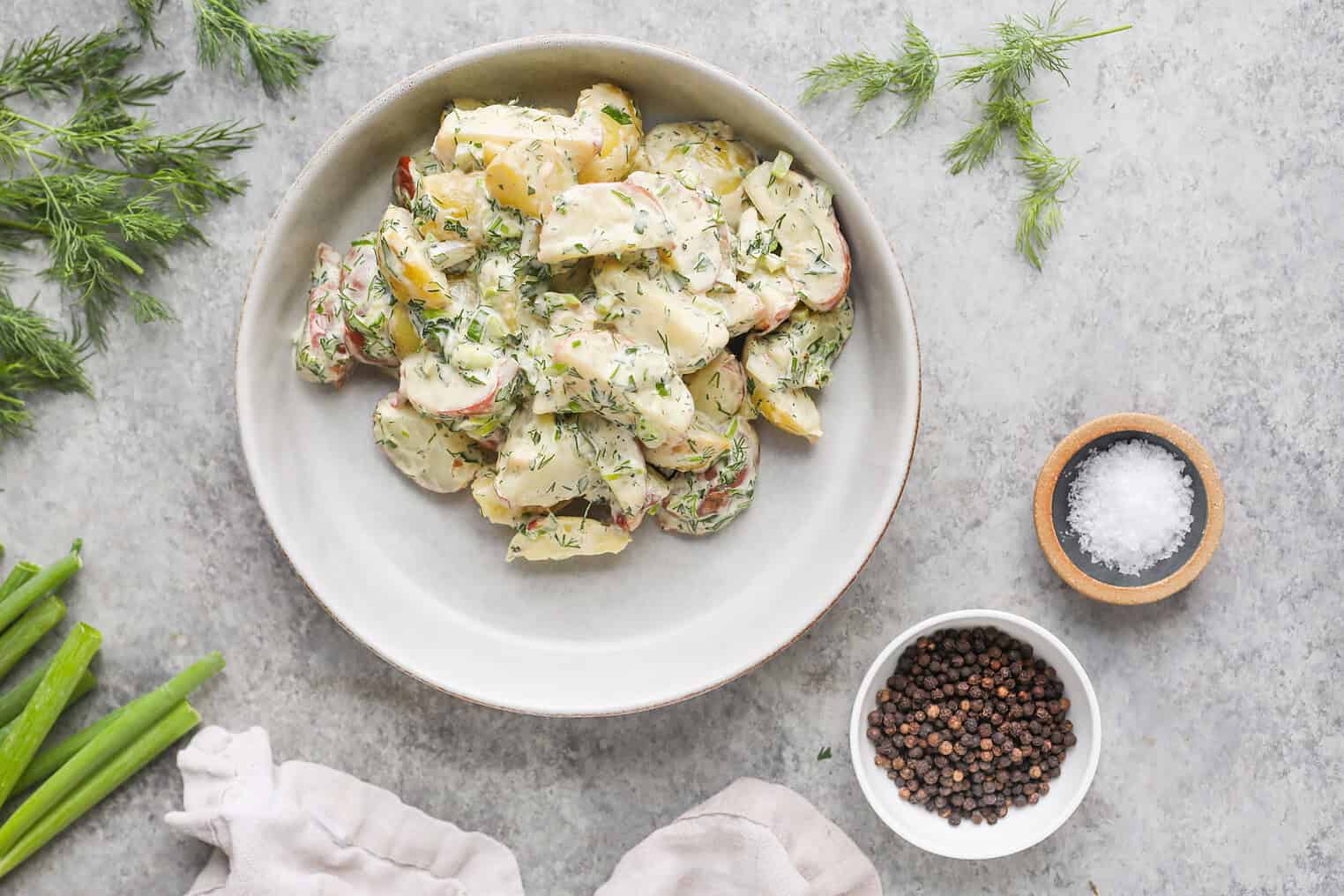 Creamy Herbed Potato Salad 2 A Bowl Of Creamy Herbed Potato Salad Garnished With Fresh Dill Sits On A Gray Surface, Surrounded By Green Onions, Dill Sprigs, A Small Bowl Of Salt, Black Peppercorns, And A Beige Cloth Napkin.