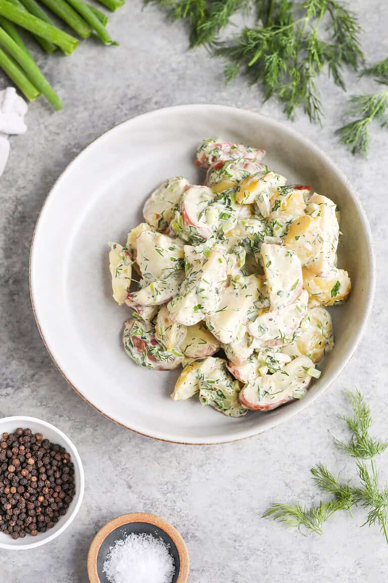 A Bowl Of Herbed Potato Salad Garnished With Chopped Dill, Surrounded By Fresh Dill, Green Onions, Black Peppercorns, And A Small Dish Of Salt On A Light Gray Surface.