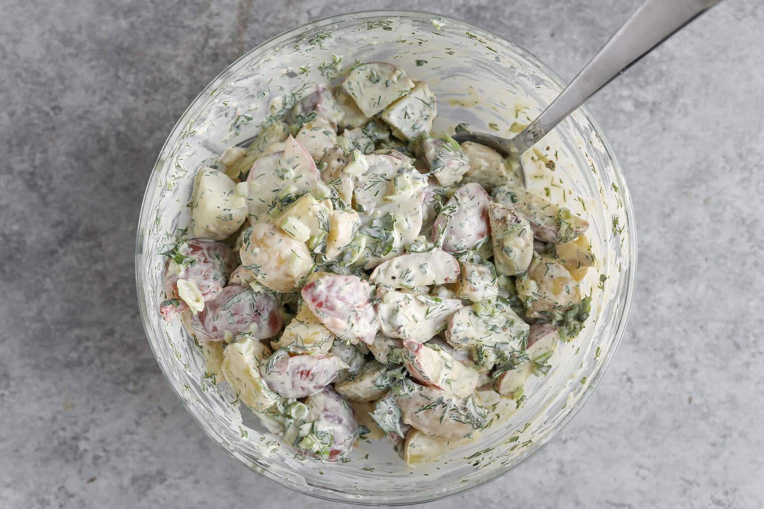 Creamy Herbed Potato Salad 8 A Glass Bowl Filled With Herbed Potato Salad, Creamy And Mixed With Chopped Fresh Herbs, With A Metal Spoon Resting Inside The Bowl, Set On A Gray Textured Surface.