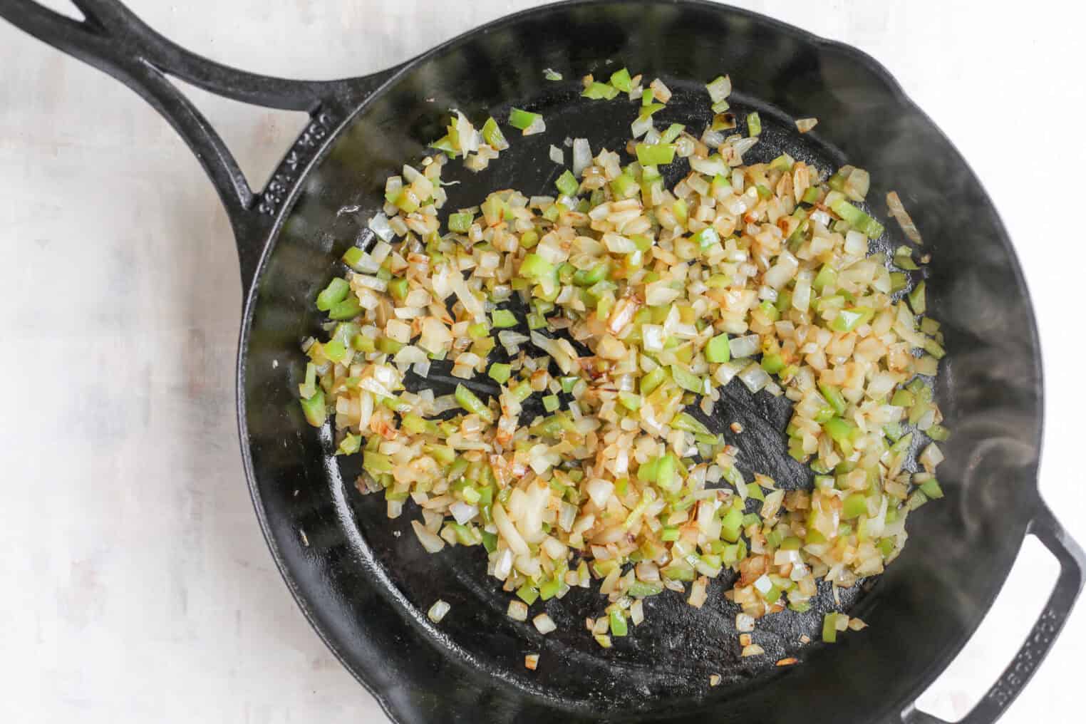 Smoky Vegetarian Baked Beans 4 Chopped Onions And Celery Are Sautéing In A Black Cast Iron Skillet, With Some Pieces Lightly Browned, Creating A Flavorful Base For Vegetarian Baked Beans, On A Light-Colored Surface.