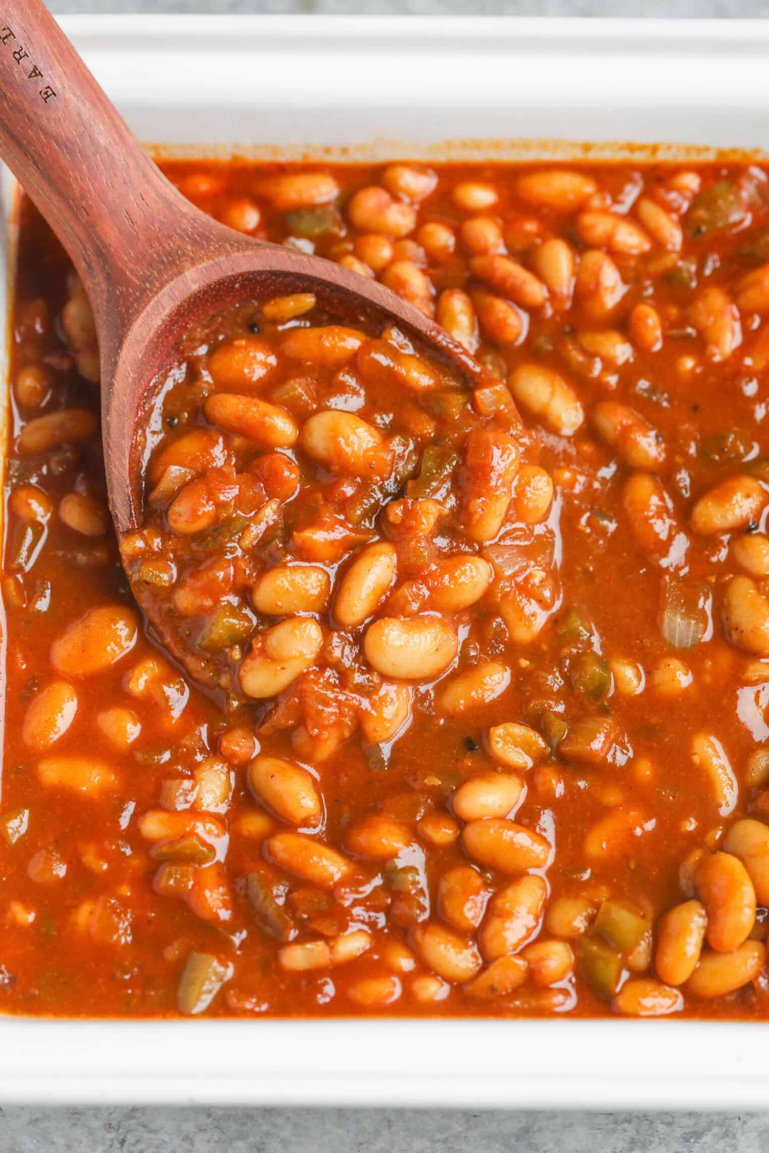 Smoky Vegetarian Baked Beans 3 A Wooden Spoon Scooping Vegetarian Baked Beans In A Rich, Tomato-Based Sauce With Visible Chunks Of Green Peppers And Onions In A White Dish.