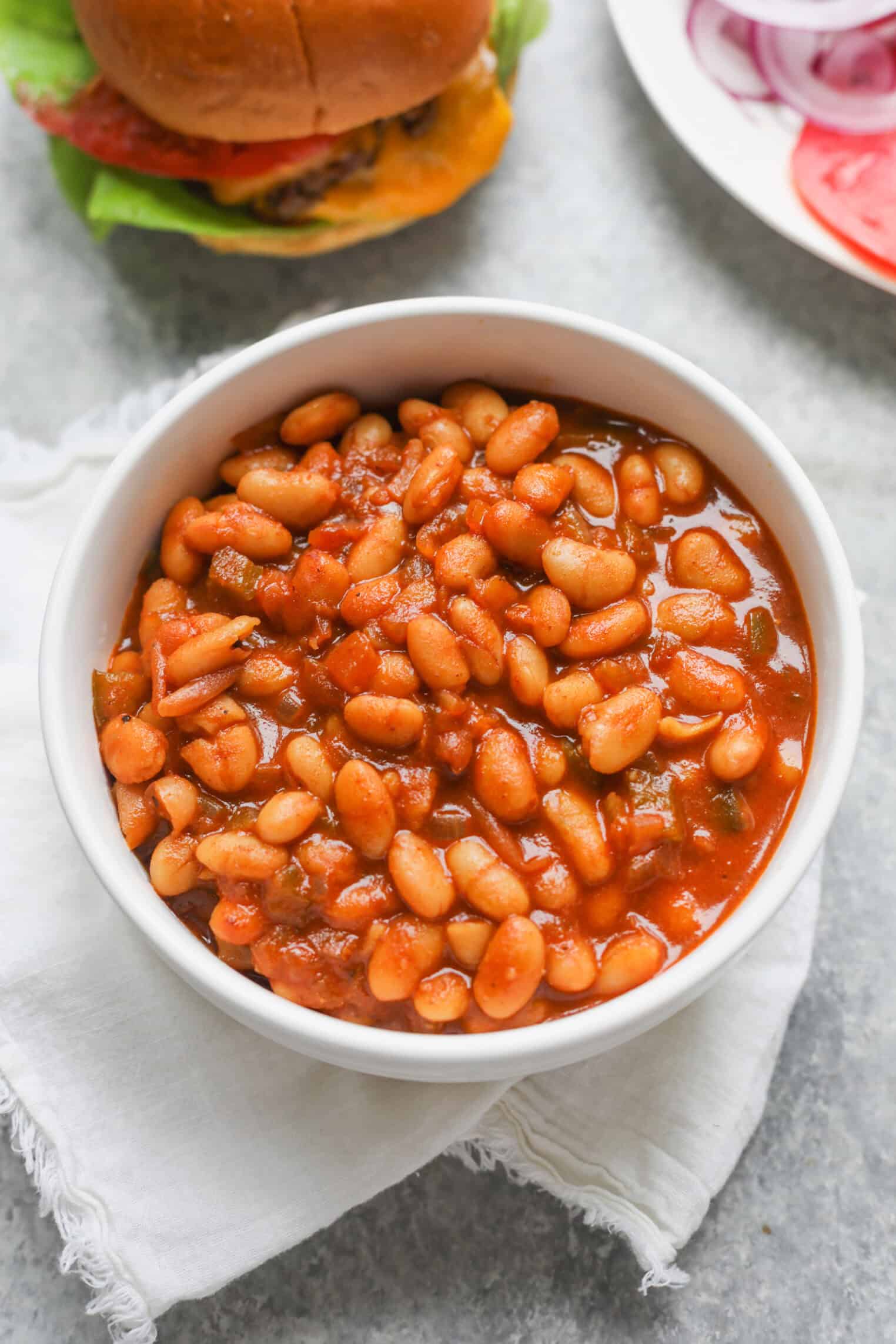 Smoky Vegetarian Baked Beans 1 A White Bowl Filled With Smoky Baked Beans In Tomato Sauce Sits On A White Cloth, With A Hamburger And A Plate Of Sliced Onions And Tomatoes Visible In The Background.