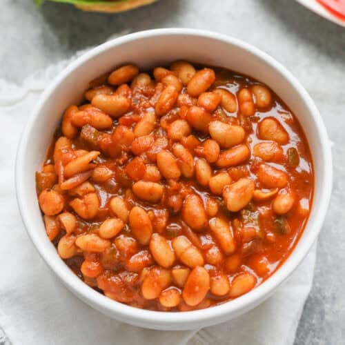 Smoky Vegetarian Baked Beans 10 A White Bowl Filled With Smoky Baked Beans In Tomato Sauce Sits On A White Cloth, With A Hamburger And A Plate Of Sliced Onions And Tomatoes Visible In The Background.