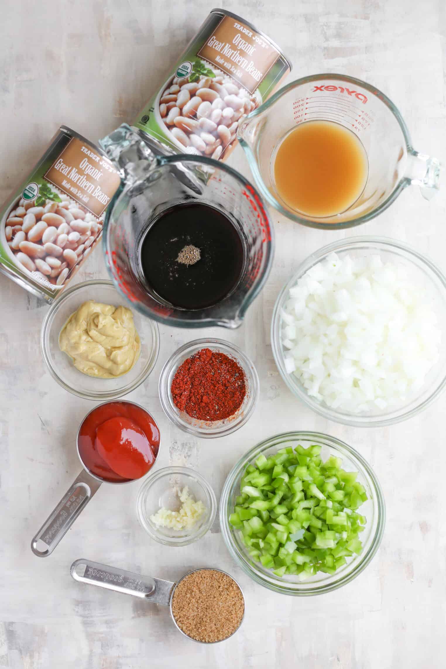 Smoky Vegetarian Baked Beans 2 An Overhead View Of Vegetarian Baked Beans Ingredients On A Light Surface, Including Two Cans Of White Beans, Chopped Onions, Chopped Celery, Tomato Sauce, Spices, Minced Garlic, Mustard, Brown Sugar, Vinegar, And Broth In Glass Bowls And Measuring Cups.